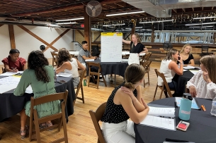 Students sit at round tables, listening to a peer present from a posterboard